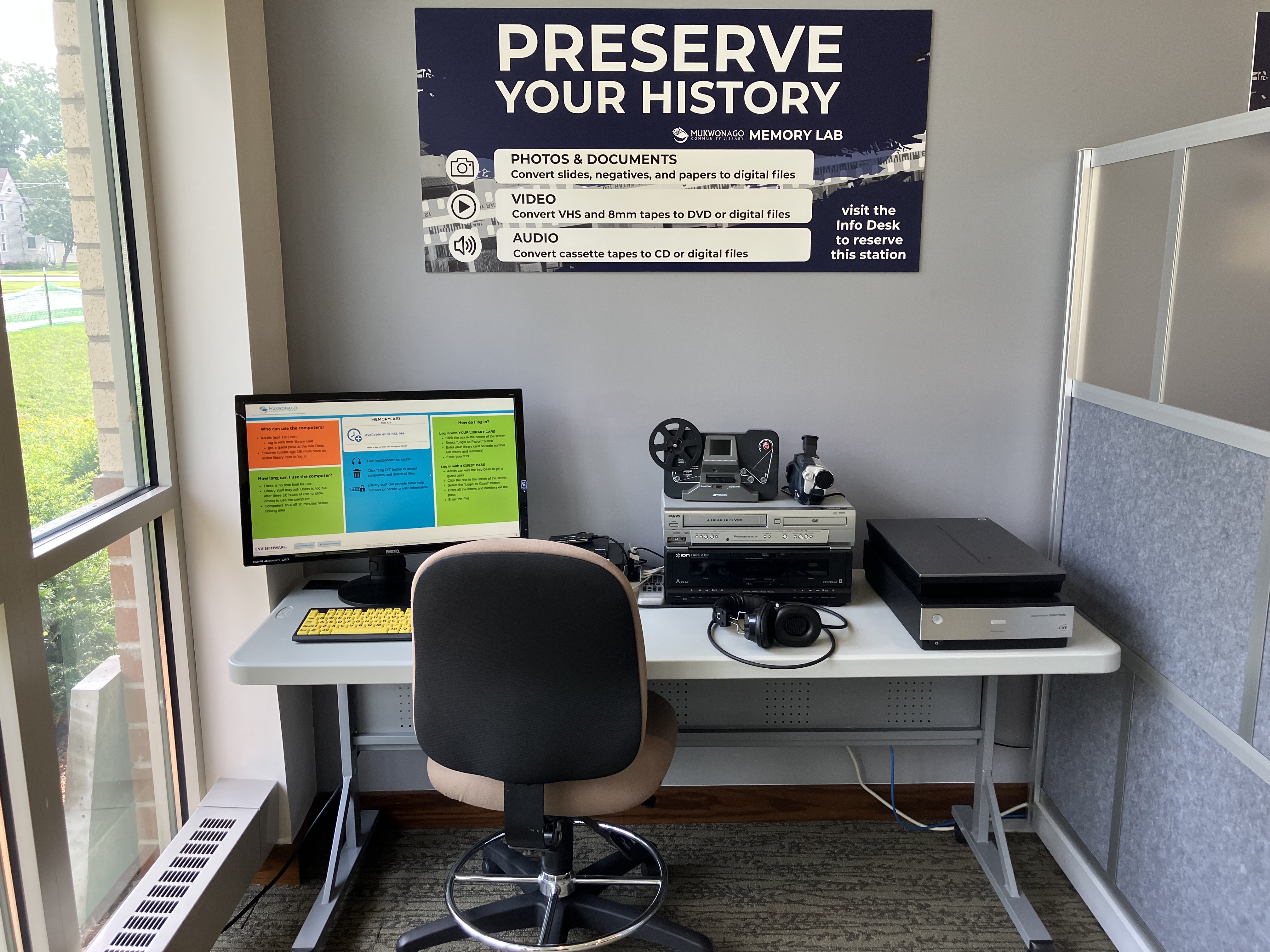 Memory Lab computer on desk with scanner, VHS player, and other digitizing equipment with sign reading "Preserve Your History"