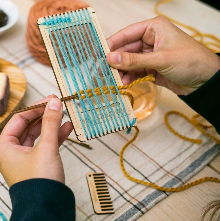 hands holding small loom with blue warp and yellow weft